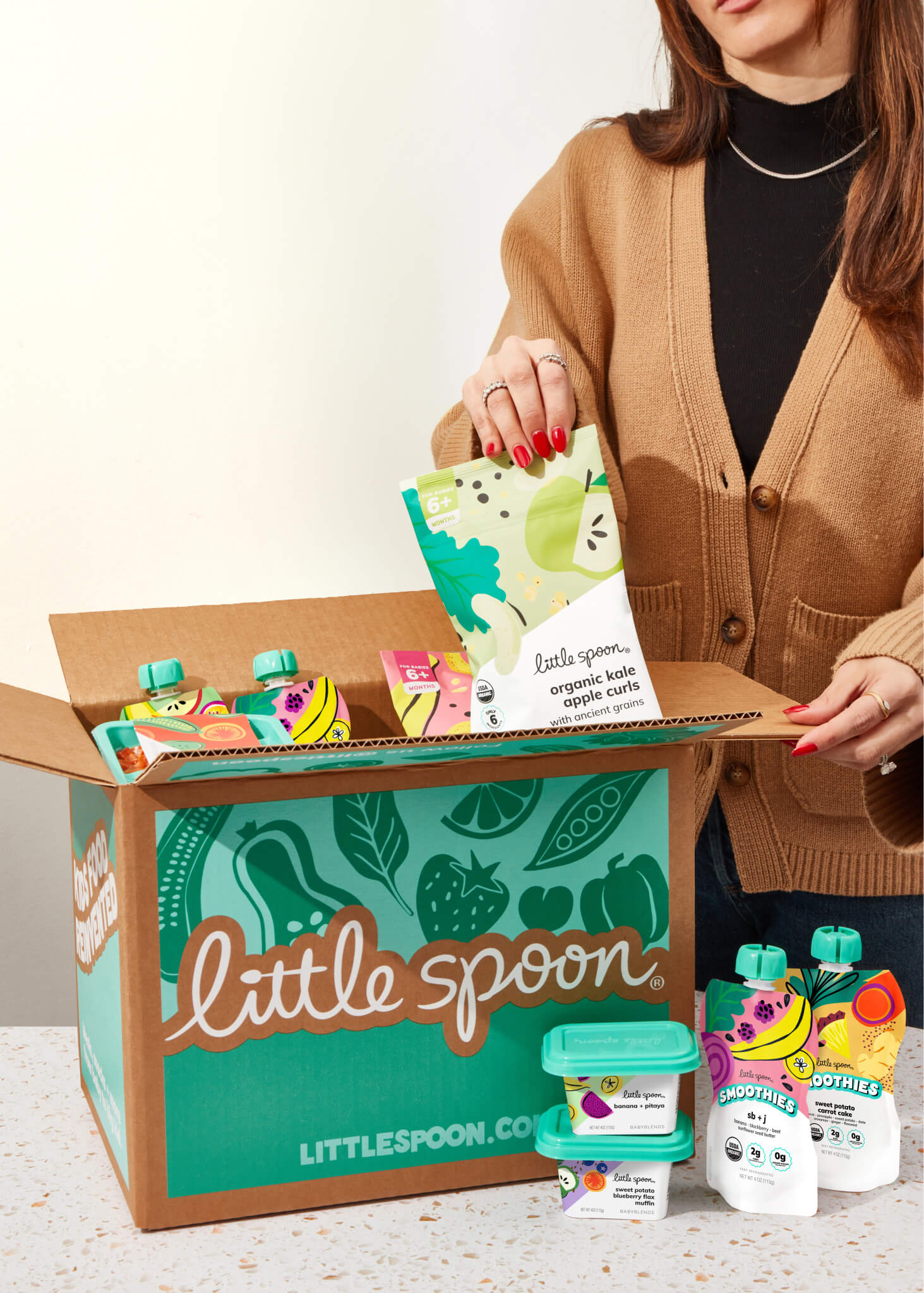 A woman unpacks a box of Little Spoon baby and kids food, holding a bag of organic kale apple curls. The box contains pouches, purees, and snacks in colorful packaging.