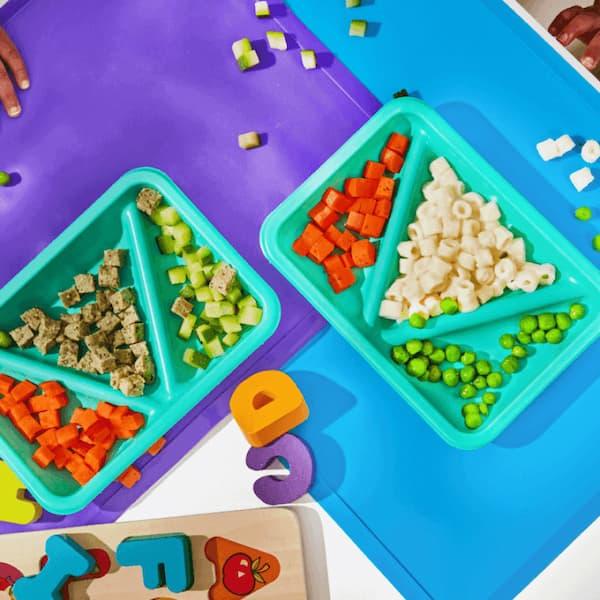 Turkey Kale Meatballs and Mini White Mac and Cheese biteables meals on purple and blue placemats with scattered food and toy letters.