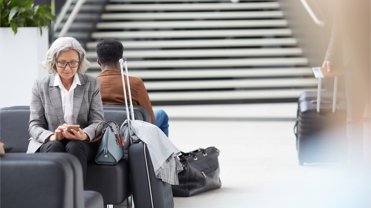 An older woman in a business suit texting on her phone at the airport with her luggage beside her.